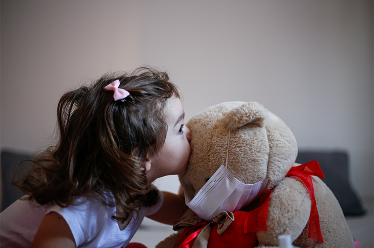 Little girl giving a kiss to a teddy bear wearing surgical mask